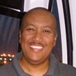 A smiling man with a shaved head, known for his keto weight loss journey, stands indoors in a gray collared shirt, with city lights glowing through a window behind him.