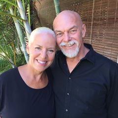 A smiling older couple with closely-cropped white hair stands together outdoors, both wearing black tops, showcasing their keto weight loss success amid greenery and a bamboo fence in the background.