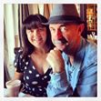 A woman with shoulder-length dark hair and bangs, in a black dress with white polka dots, sits beside a man in a gray fedora and blue shirt. Smiling over drinks indoors, they chat about keto weight loss successes.