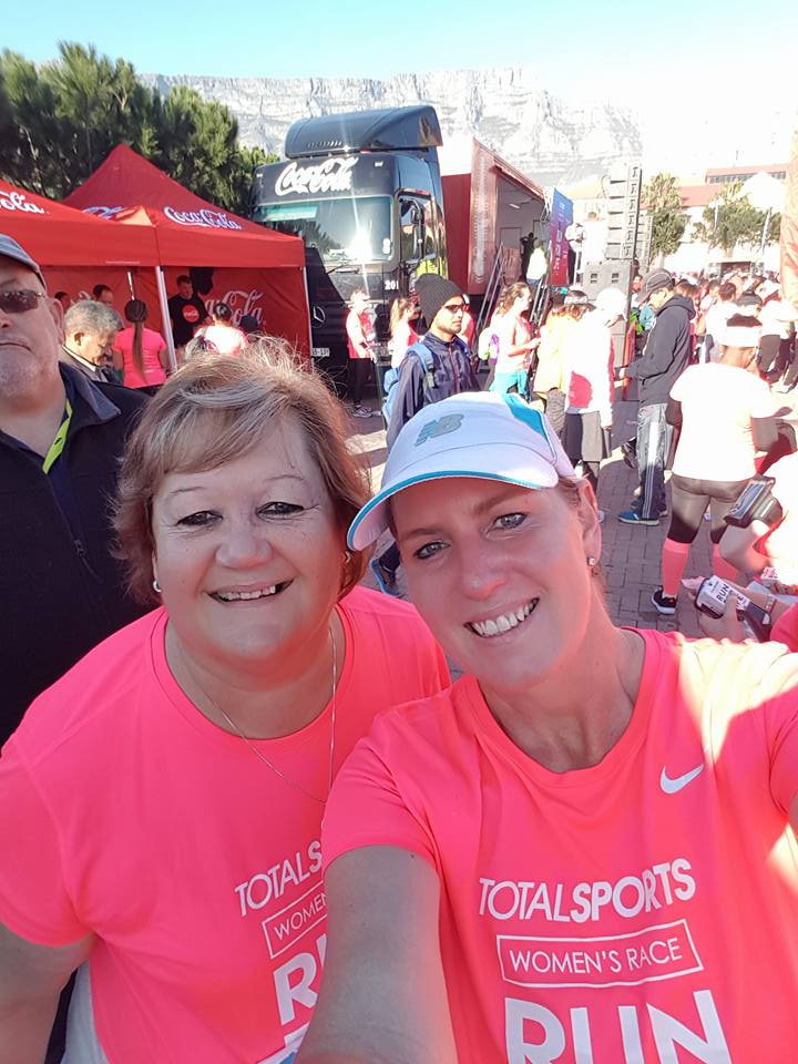 Two women wearing bright pink Totalsports Womens Race shirts smile for a selfie at an outdoor event, celebrating their active lifestyle and keto weight loss journeys. Other participants and tents are visible in the sunny background.