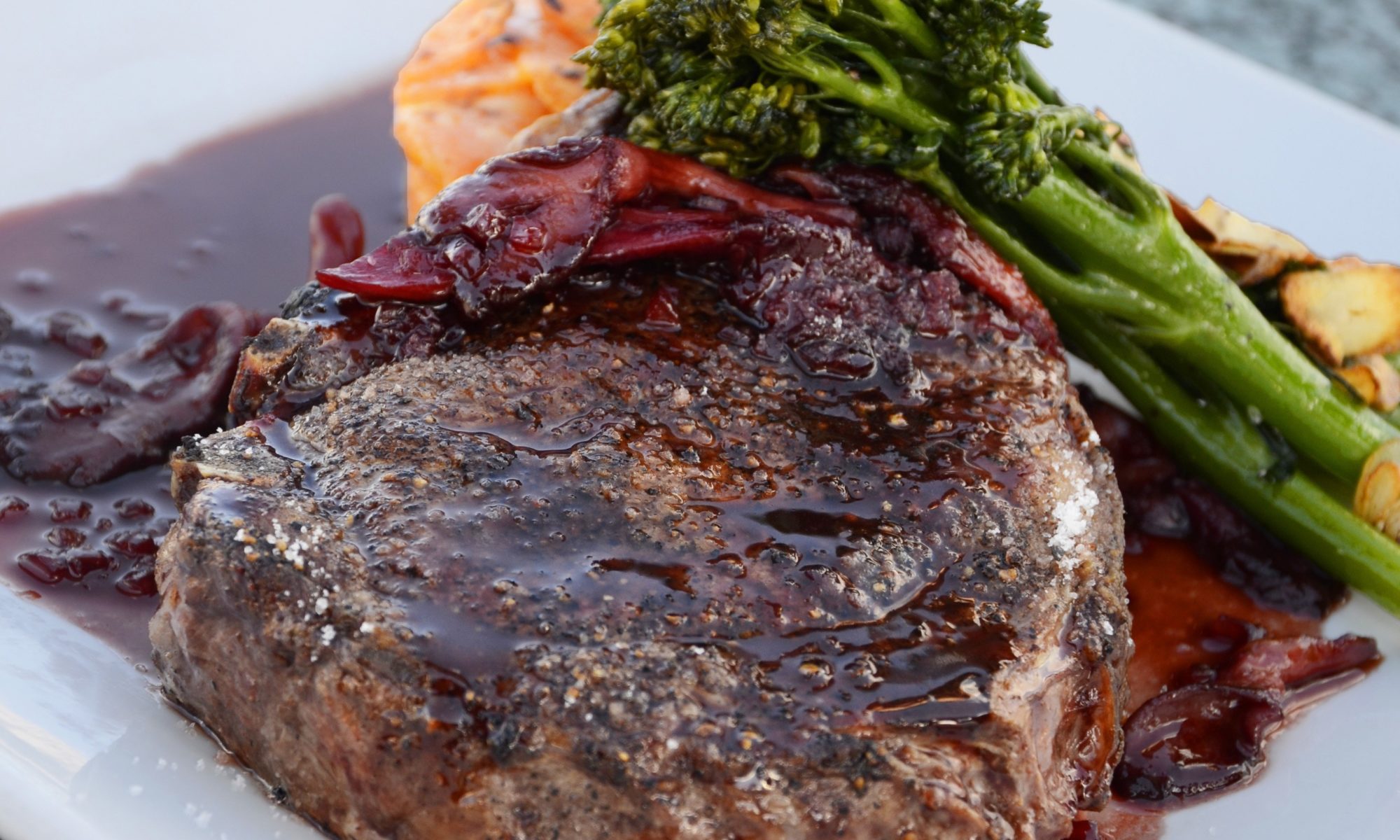 A cooked steak topped with a rich, dark sauce sits on a white plate, accompanied by broccolini and sliced roasted potatoes—perfect for those exploring keto weight loss—plus a small serving of cheesy pasta in the background.