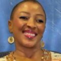 A woman with short hair smiles warmly at the camera, radiating confidence in her keto weight loss journey. She wears large circular earrings, a beaded necklace, and glossy pink lipstick. The background is blue.