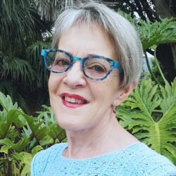 A smiling older woman with short gray hair and blue glasses stands outdoors in front of large green tropical leaves, showcasing her vibrant spirit and the confidence she gained through keto weight loss, wearing a light blue textured top.